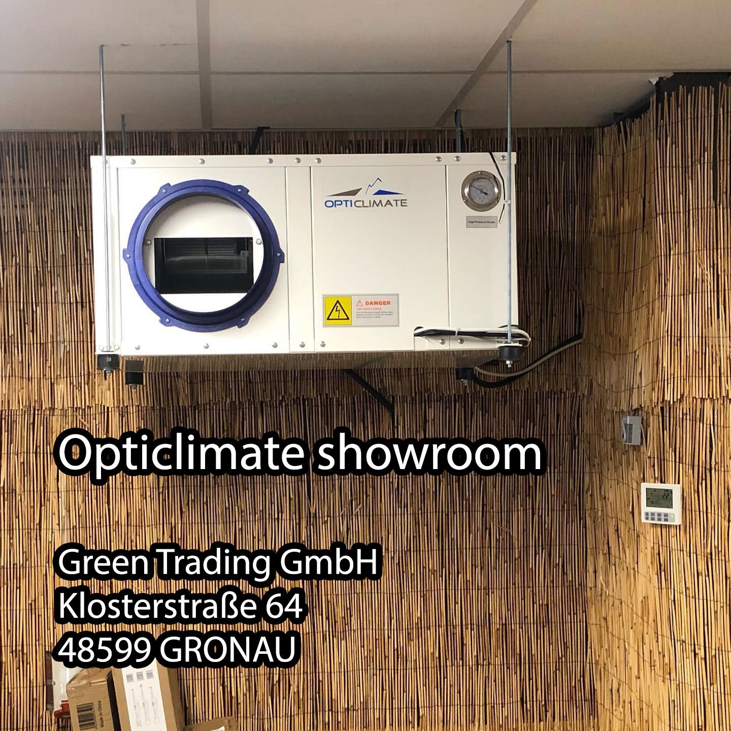 Opticlimate systeem bevestigd aan een wand in het showroom van Green Trading GmbH. Het systeem heeft een witte behuizing met een blauwe rand om de luchtinlaat. Aan de voorkant is een manometer zichtbaar en er zijn waarschuwingslabels aangebracht. De achtergrond heeft een natuurlijke uitstraling door het gebruik van bamboe panelen. Het systeem is ontworpen voor klimaatbeheersing in kassen en andere teeltomgevingen, met aandacht voor energie-efficiëntie en duurzaamheid.